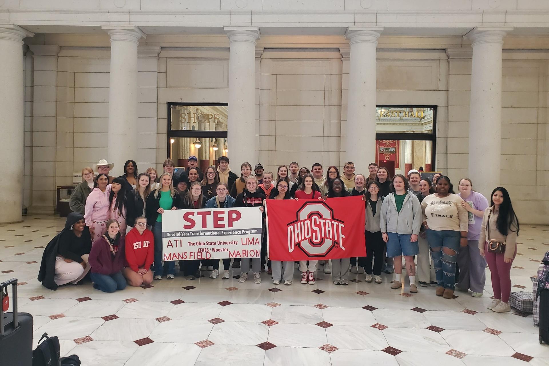 large group of students in front of ornate columns holding banner and flag