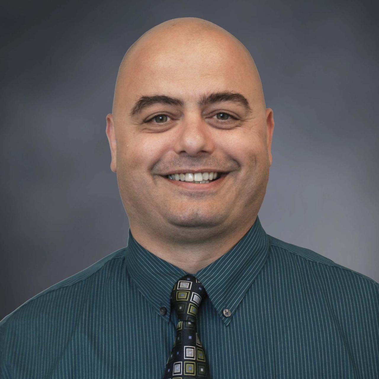 Head and shoulders of a bald man smiling in bluish green shirt and matching tie