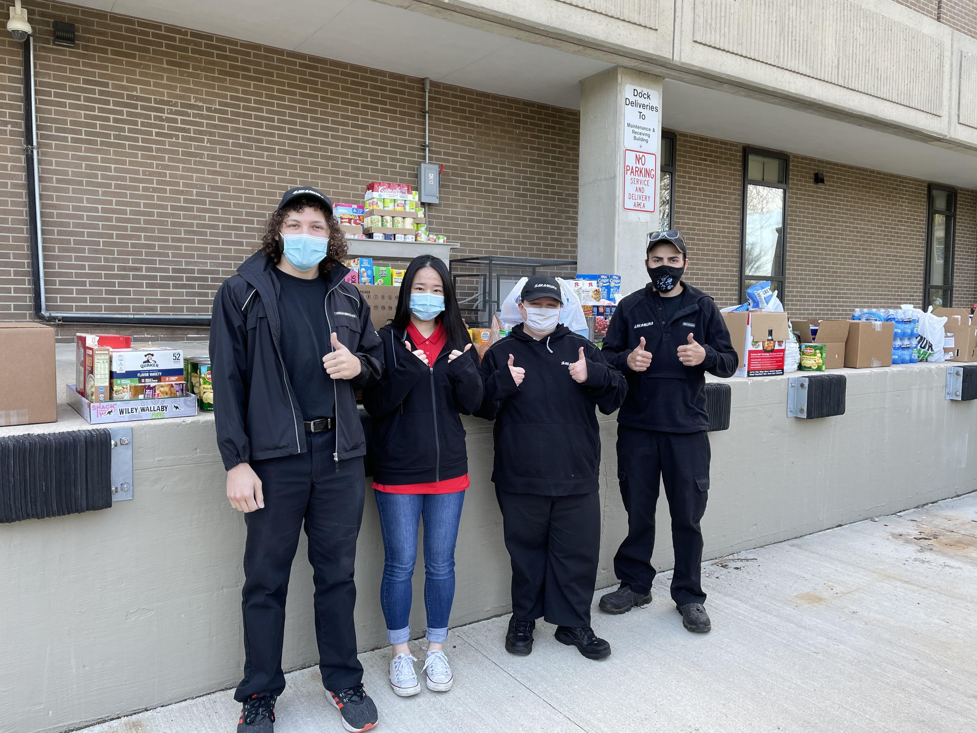 Group of 4 dressed in black stand in front of loading dock of food with thumbs up