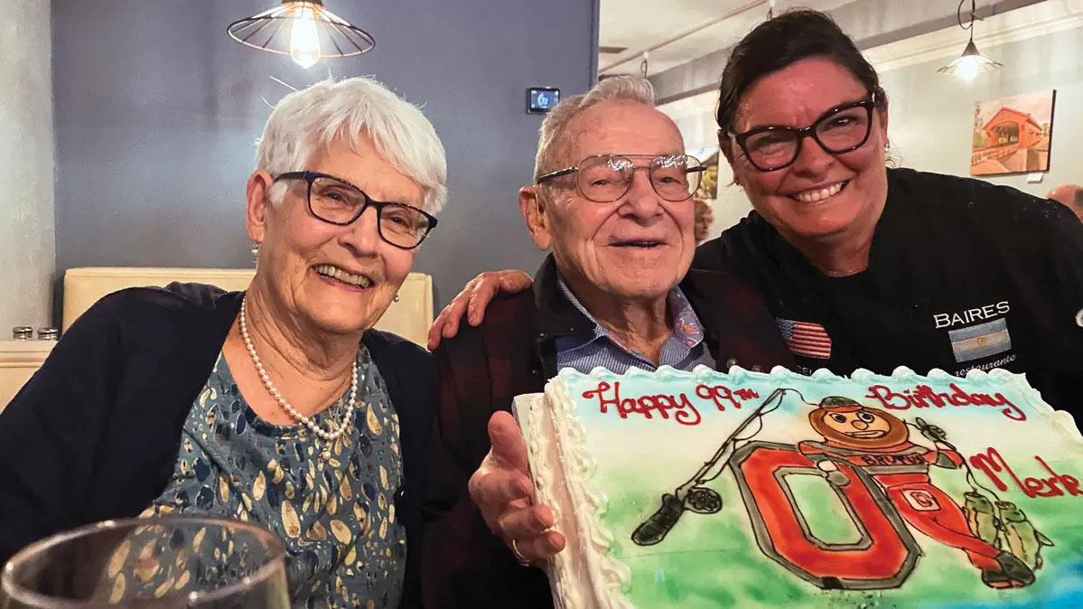 Older man in glasses with birthday cake surrounded by two smiling women