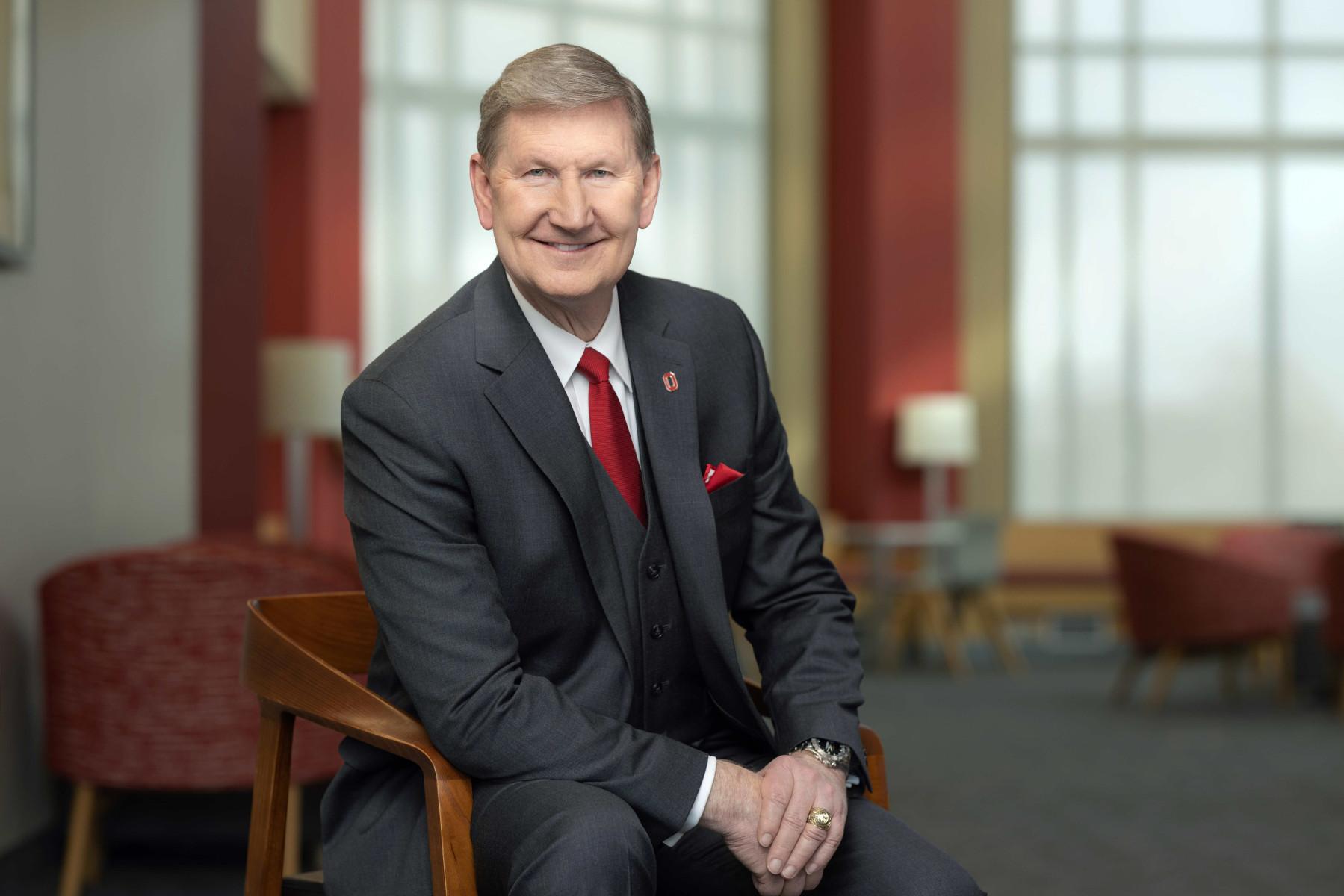 man in grey suit, red tie, and handkerchief seated smiling with windows behind