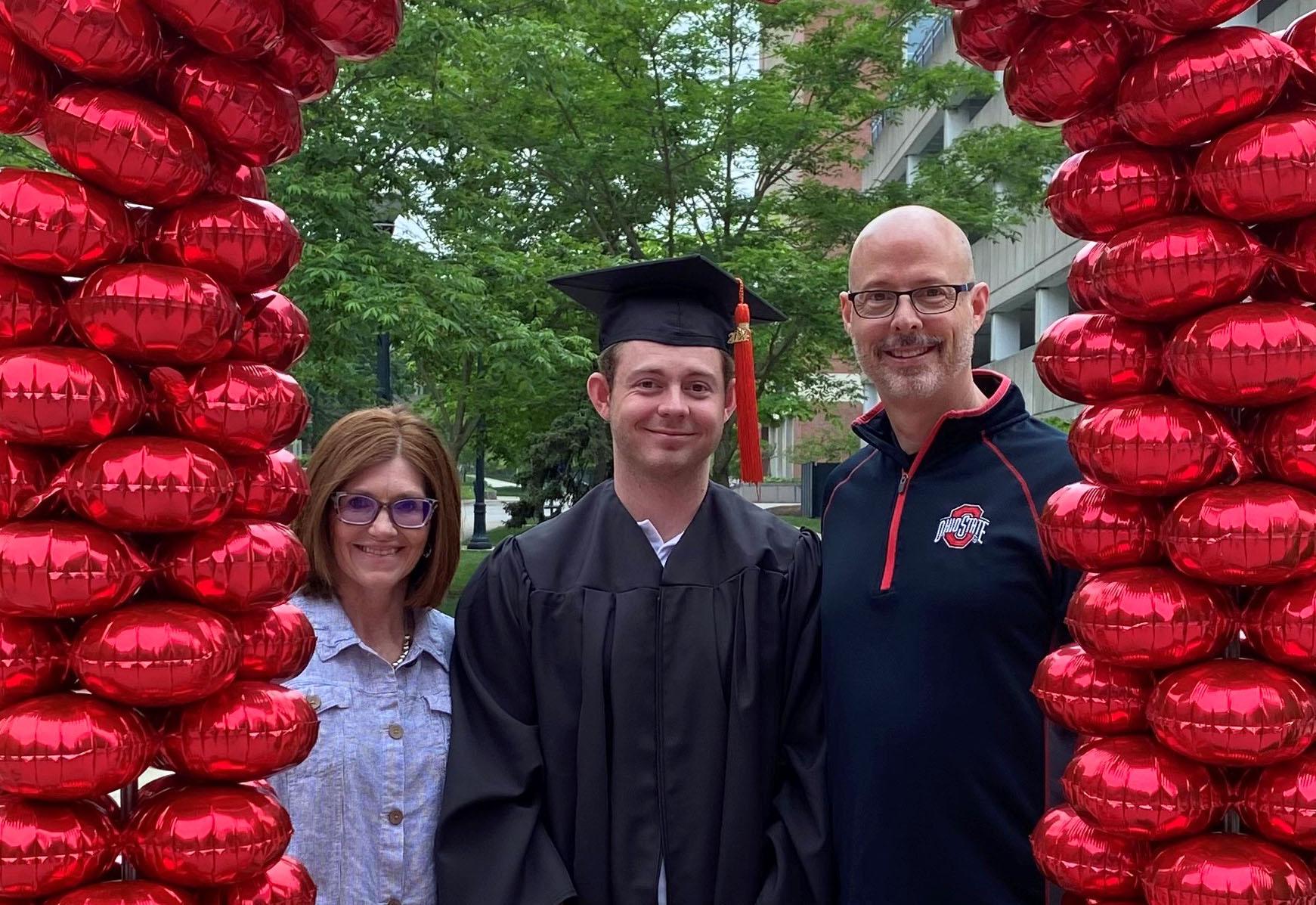 graduate in cap and gown with parents surrounded by red balloons 