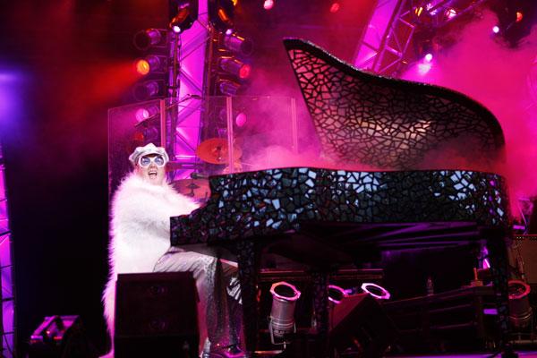 man with stage lights and elton john costume performing behind grand piano