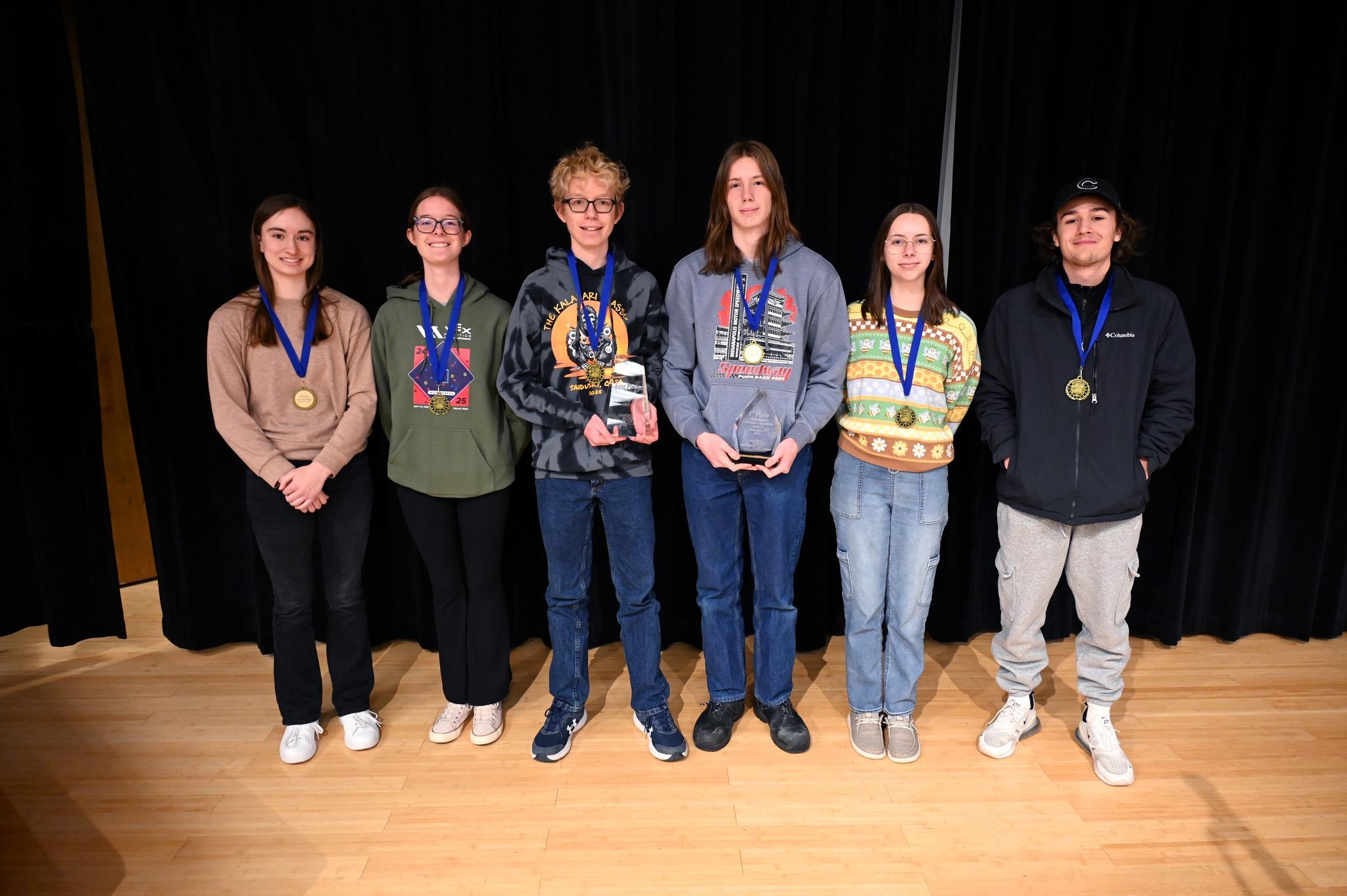 Six male and female students on stage holding a trophy and wearing medals