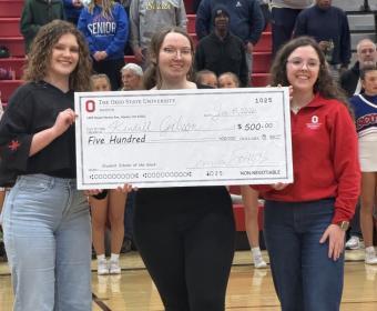 Young woman receives large check at basketball game from 2 other women