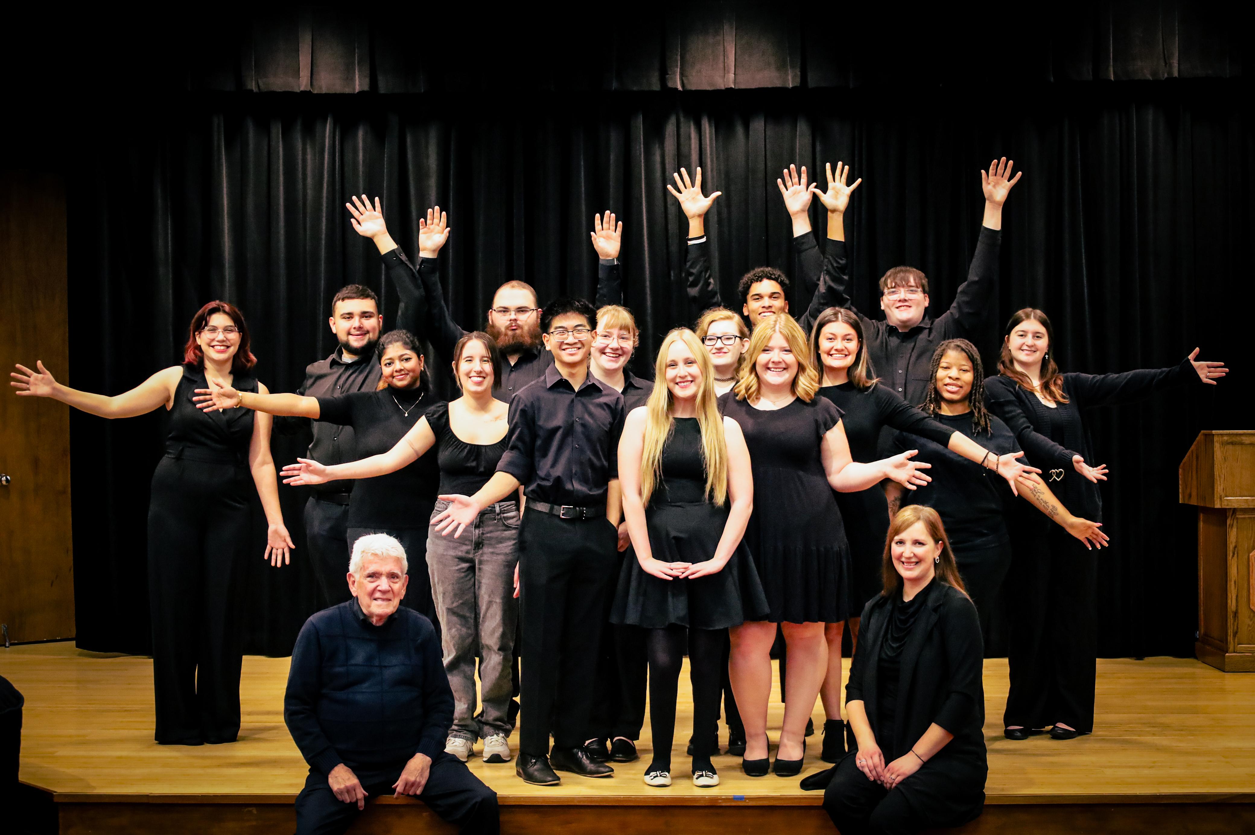 singing group posed on stage with arms outreached dressed in all black