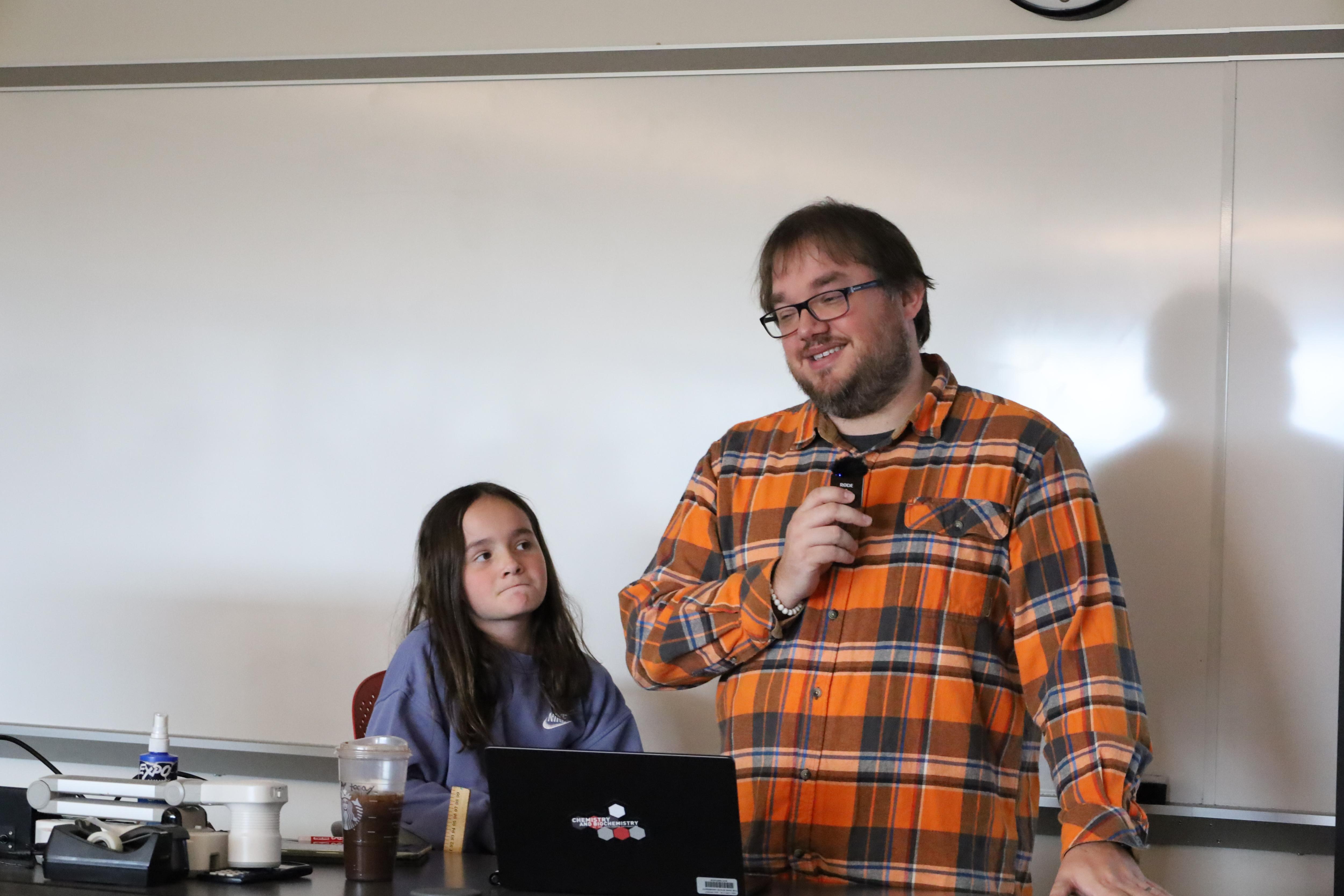 bearded man in front of white board standing next to young daughter speaking to a crowd