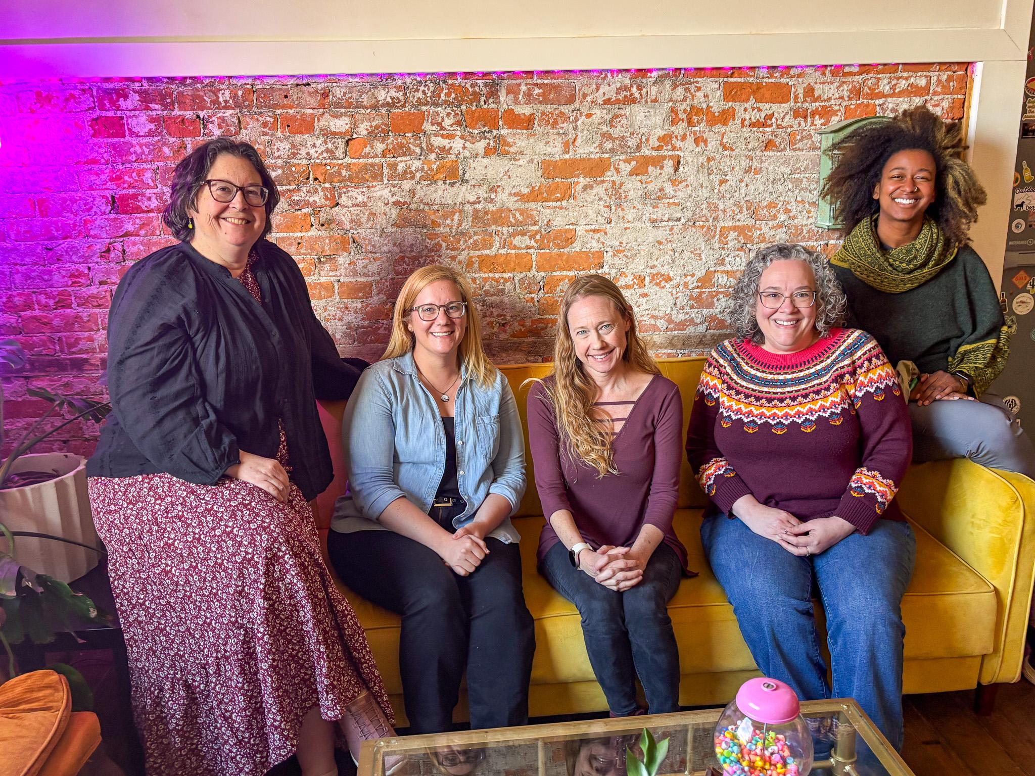 Five women seated at yellow coach with brick wall in the background