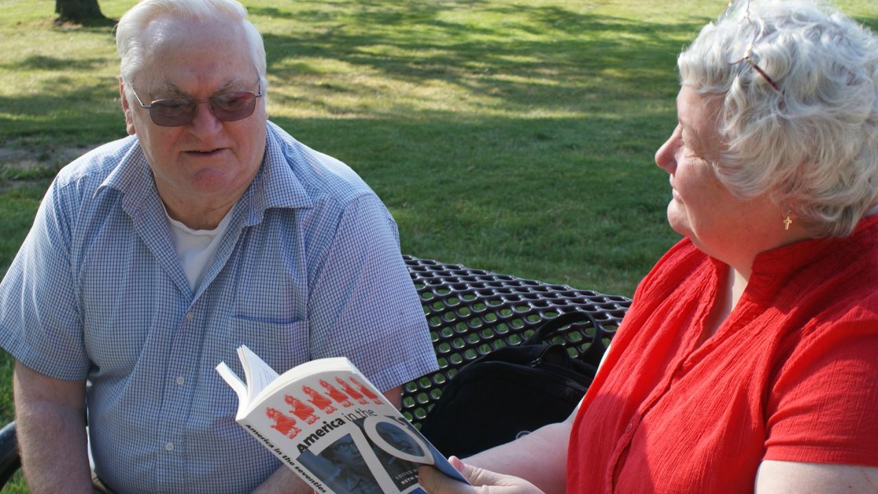 older woman reads from text with older man on bench