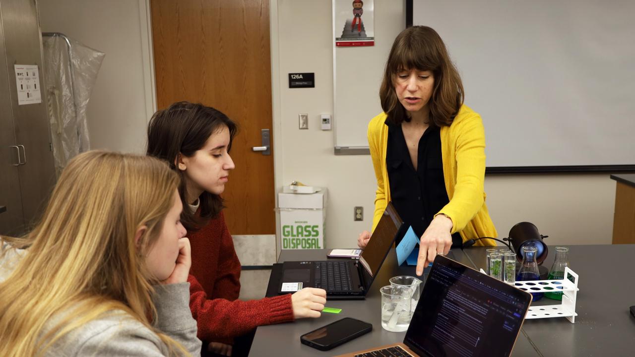 women teaching two students at laptops