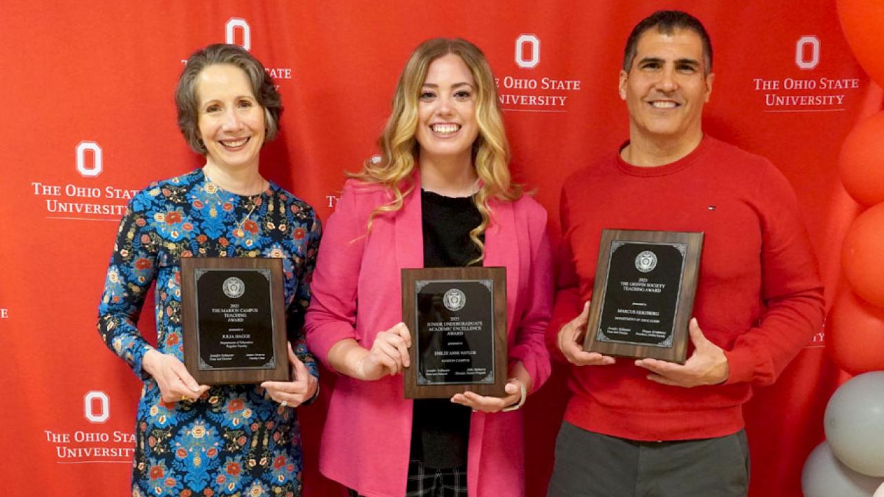 Two women and a man holding plaques in front of red logo backdrop