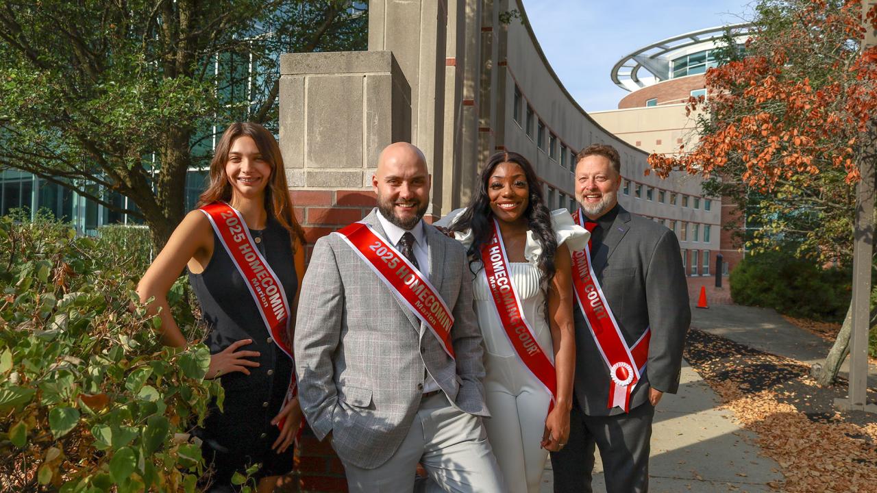 two women and two men in dress clothes and sashes in front of campus library